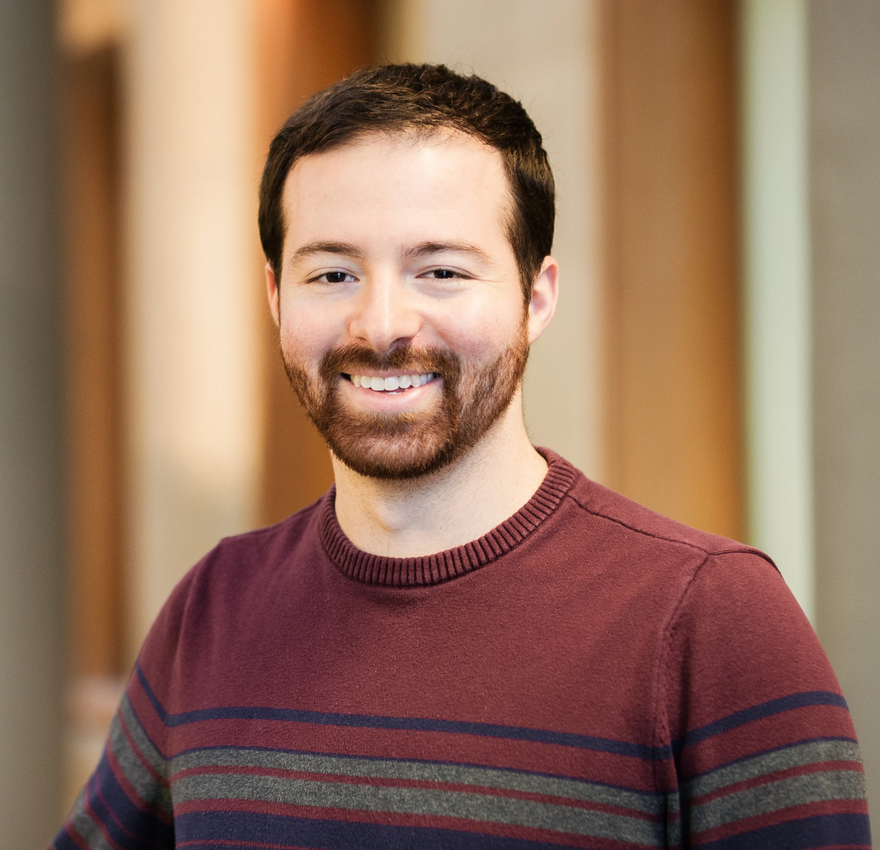 Headshot of a man from the chest up. His hair is brown and he has facial hair. He is smiling at the camera wearing a…