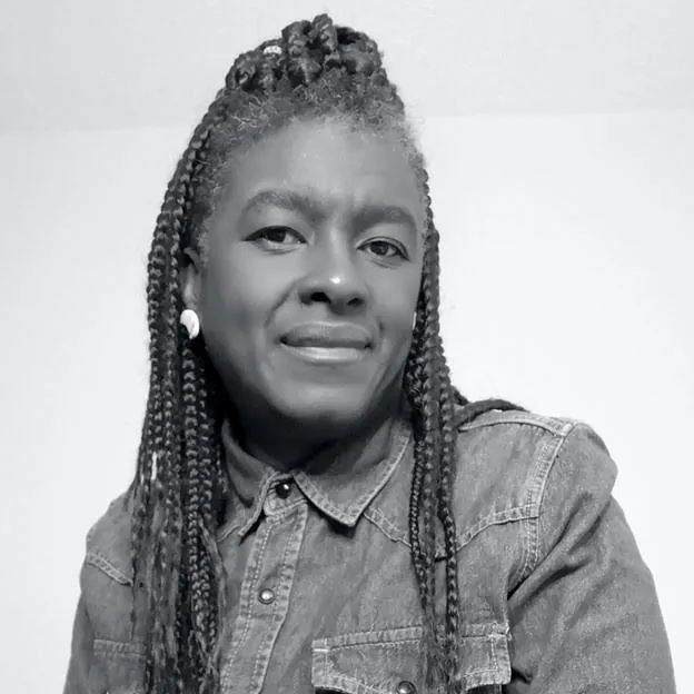 Black and white headshot of a Black woman, Jo-Anne, who has long braided hair and is wearing earrings. She is smiling at the…
