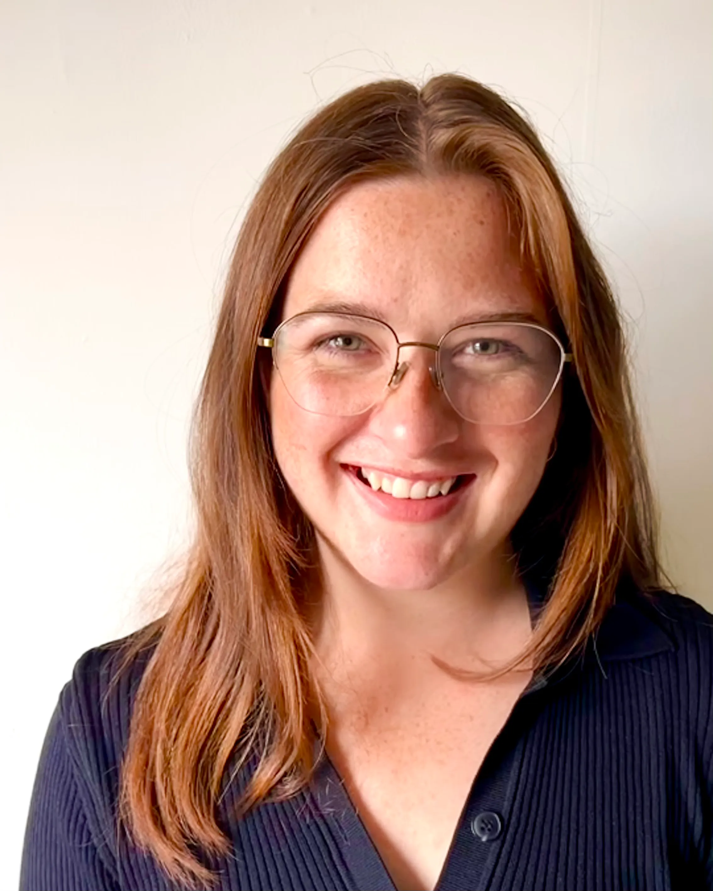 Image of a white woman smiling at the camera. She has long red hair parted in the middle, copper-framed glasses, and is…