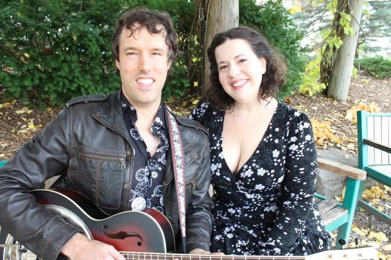 A photo of musicians Luke Haneman and Meghan Lawrence. Luke has short dark wavy hair, wears a patterned shirt and black…