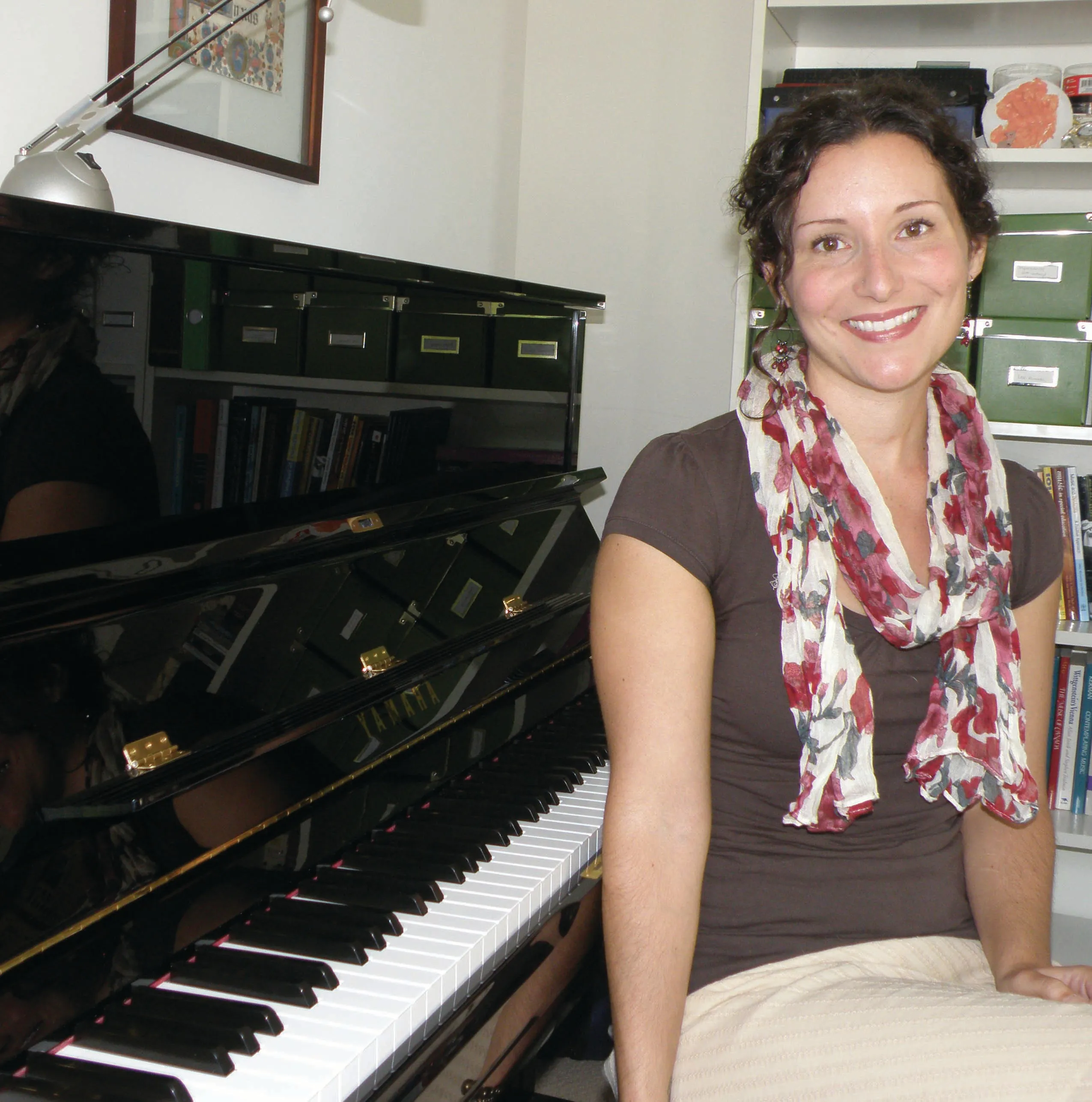 An image of Erin sitting to the right of an upright piano. She is wearing a flower-patterned scarf and is smiling at the…