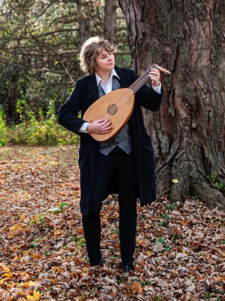 Rowan Friesen stands in a forest in autumn wearing a black suit with silk vest and white open collar shirt. He has wavy…