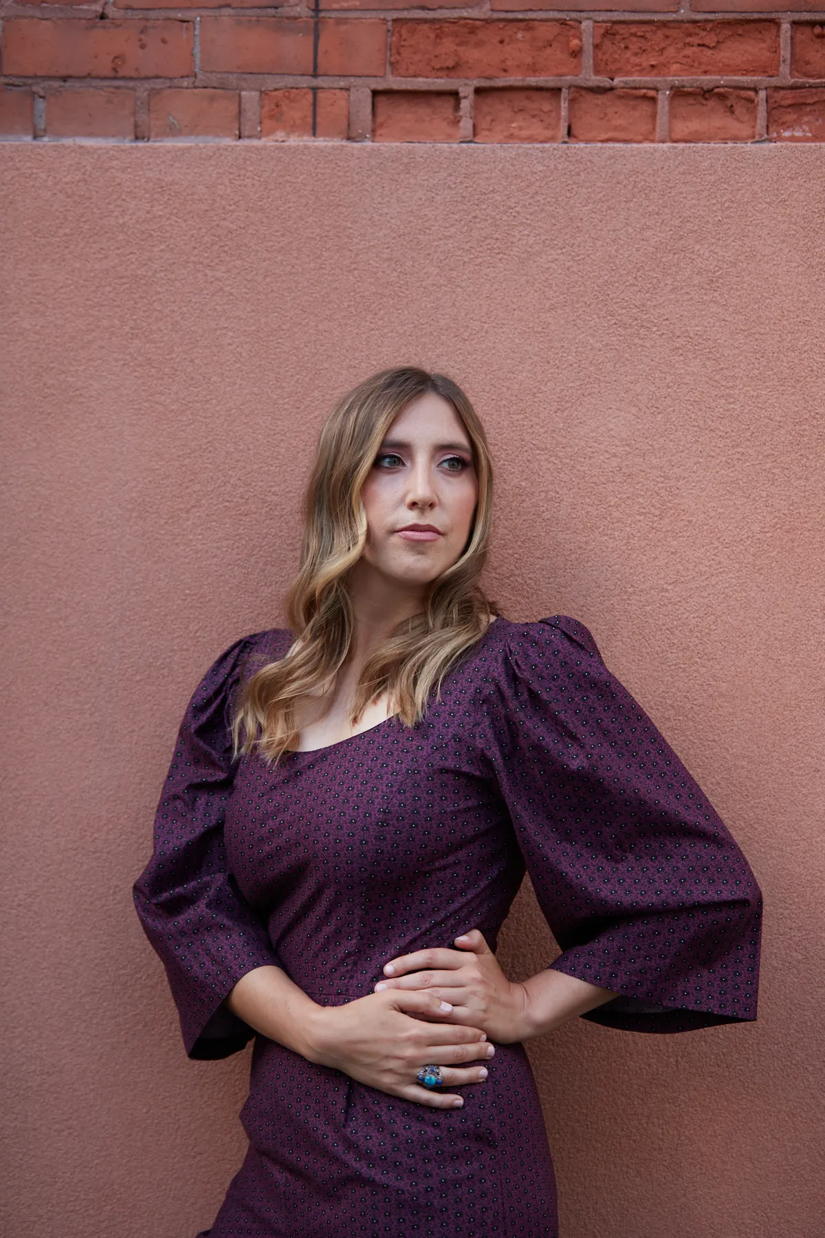 Headshot of a woman. She has long, flowy light brown hair parted in the middle, and is wearing a purple, patterned dress.…