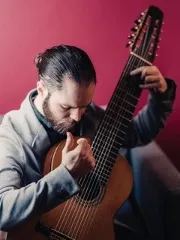 Julian has long brown hair tied up in a bun, a long beard, and white skin. He is holding a guitar with 10-strings and…