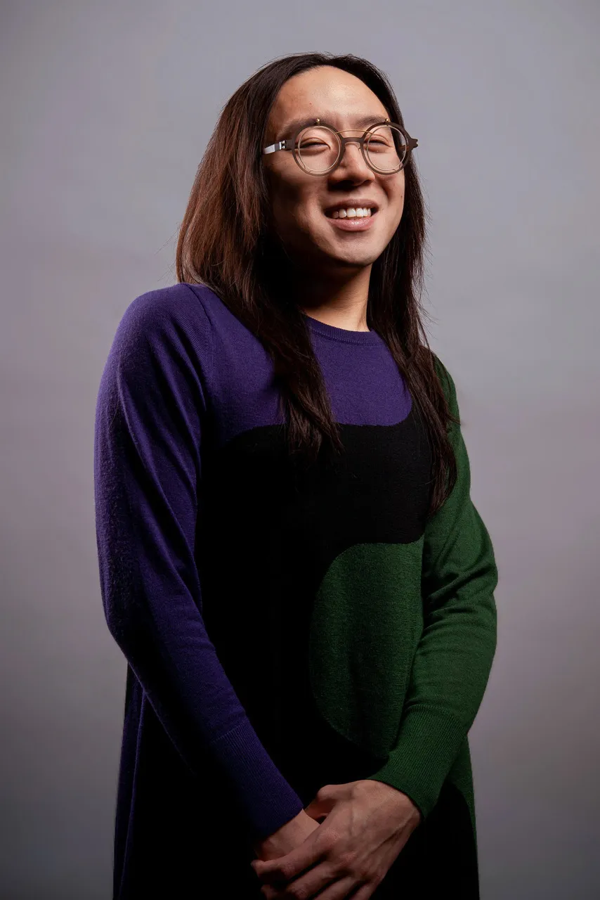 Sean Lee, a male-presenting East-Asian Chinese person smiles at the camera. He wears steampunk circle glasses that are brown…