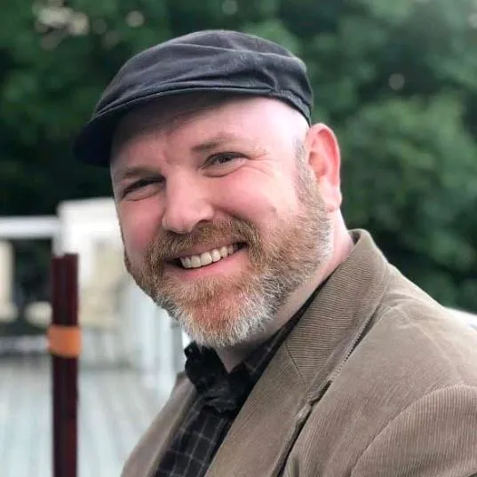 Image of a man outside smiling at the camera. He has facial hair, and is wearing a dark brimmed hat, a plaid button-up…