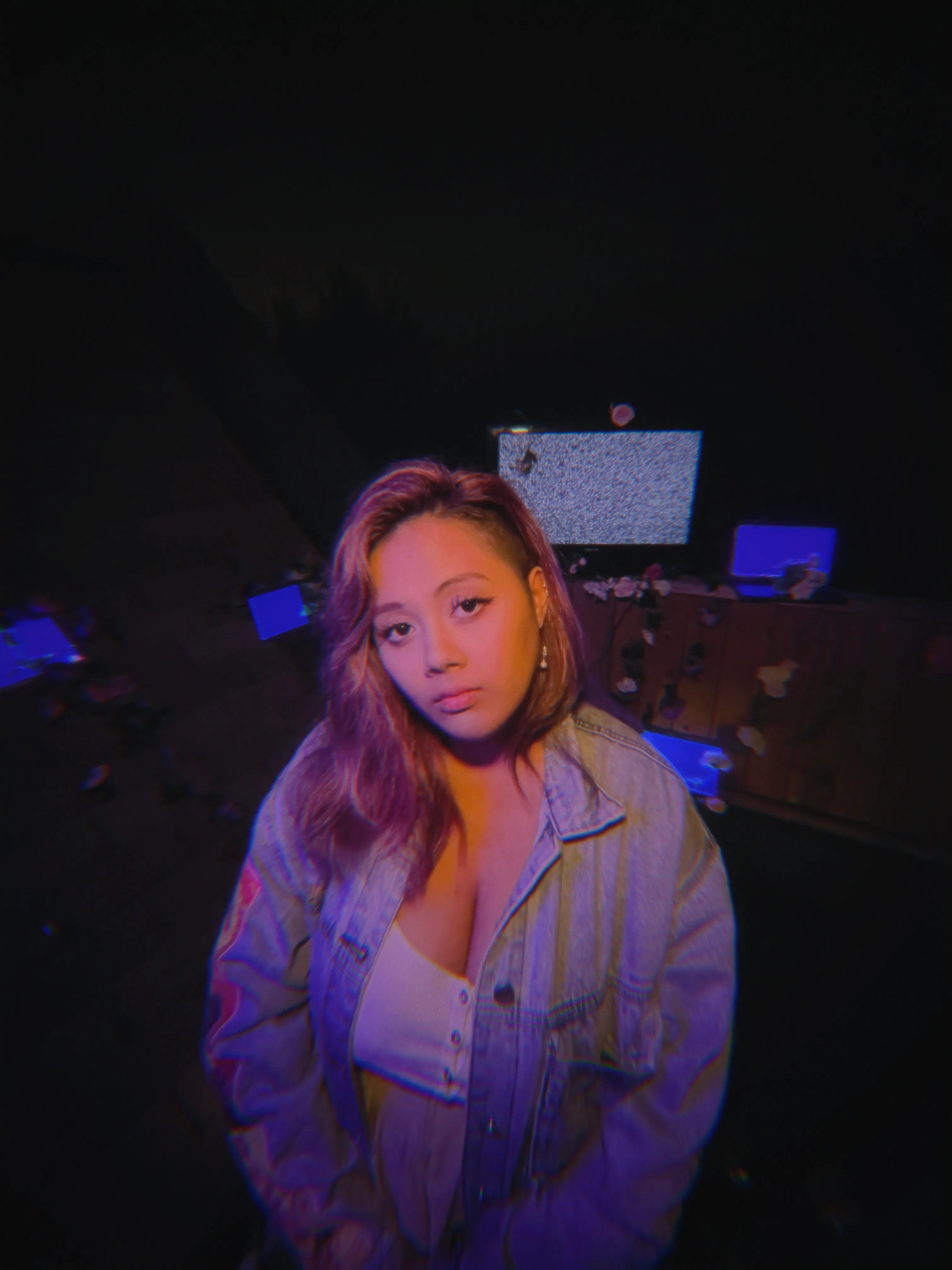 An asian woman with light pink hair "Dani Saldo", stands in the foreground wearing a denim jacket with orange patches and a…